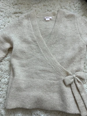 Beige omlottkofta från Gina Tricot - Mysig beige omlottkofta från Gina Tricot i mjuk stickad kvalitet. Koftan har v-ringning, långa ärmar och knytdetalj i sidan som ger en snygg siluett. Perfekt för lager på lager under höst och vinter. Passar perfekt till jeans/kostymbyxor eller kjol. Super mysig och perfekt för att ”toucha” upp outfiten lite. Denna har används max 2 ggr så nästan helt ny!❣️❣️