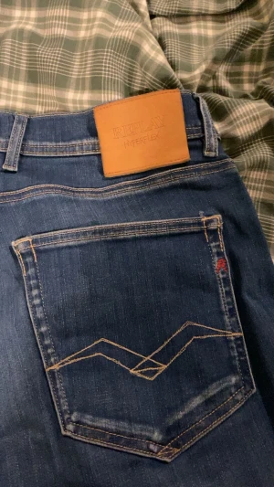 Blå Replay Hyperflex Grover jeans - Säljer ett par blå Replay Hyperflex Grover jeans med klassisk femficksdesign och snygga kontrastsömmar på bakfickorna. Jeansen har en skön stretchig känsla och normal passform. Märkespatch i läder baktill i midjan och diskret Replay-logga vid fickan.