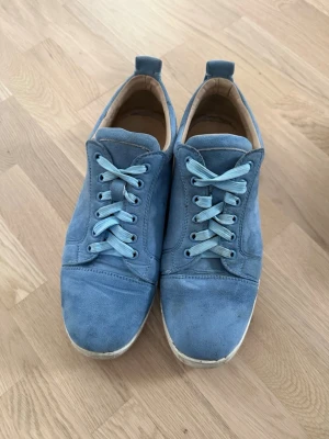 Christian Louboutin  - Säljer ett par stilrena blå sneakers från Christian Louboutin i mjuk mocka. Skorna har ljusblå snörning, rund tå och vit sula. Insidan är fodrad i beige skinn för extra komfort. Perfekta för dig som gillar klassisk men avslappnad stil.