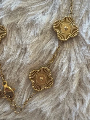 Guldig armband i rostfritt stål - Snyggt armband i guldtonad metall med fem blommor som har räfflade detaljer och pärlformad mitt. Kedjan är tunn och har ett smidigt lås. Perfekt accessoar för att lyfta din outfit med en touch av blommig elegans.