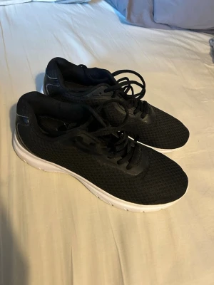 Svarta sneakers med vit sula - Säljer ett par stilrena svarta sneakers med vit sula. Ovandelen är i mesh-liknande syntetmaterial som andas och skorna har klassisk snörning. Perfekta till vardags för dig som gillar enkel och sportig stil.