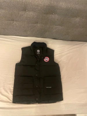 Canada Goose väst tar emot byten.  - Säljer en svart dunväst från Canada Goose med hög krage och klassisk logga på bröstet. Västen har tryckknappar framtill, två stora fickor och diskret Canada Goose-märke på ena fickan. Perfekt för lager-på-lager under kyliga dagar.