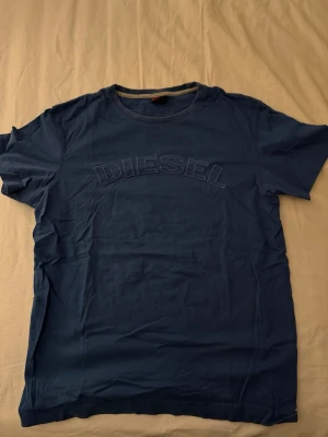 Blå t-shirt - Diesel - Snygg blå t-shirt från Diesel i storlek L med broderad logga framtill. Klassisk rund hals och korta ärmar. Tillverkad i mjuk bomull som är skön mot huden. Perfekt för dig som gillar stilrena och coola plagg med diskret branding.