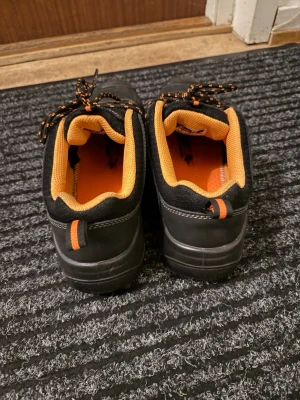 Svart/oranga jobbskor  - Säljer ett par stabila jobbskor/syddsskor i svart med orange detaljer. Skorna är gjorda för arbete inom t.ex. lager, industri, verkstad eller serviceyrken där man står och går mycket.