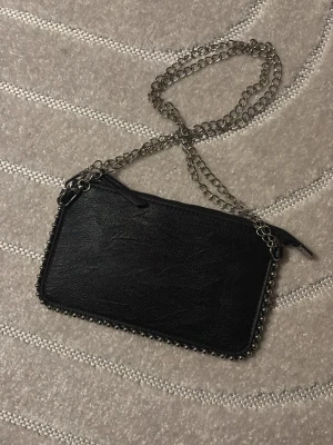 Svart axelväska med kedja - Säljer en svart axelväska i skinnimitation med silverfärgad kedja som axelband. Väskan har dekorativa små nitar längs kanterna och stängs med dragkedja. Perfekt storlek för det viktigaste och ger en cool vibe till din outfit.