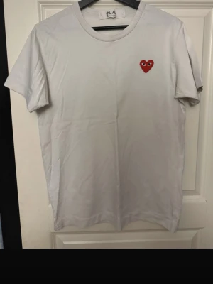 Vit t-shirt med rött hjärta från PLAY (cdg) - Vit t-shirt från PLAY med ett broderat rött hjärta med ögon på bröstet. Klassisk rund hals och korta ärmar. Tillverkad i mjuk bomull som känns skön mot huden. Perfekt för dig som gillar stilrena plagg med en twist.