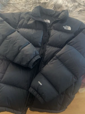 Svart pufferjacka från The North Face - Säljer en svart pufferjacka från The North Face, modell 700. Jackan har hög krage, dragkedja framtill och klassisk logga på bröstet och ryggen. Fylld med dun och perfekt för kalla dagar. Enkel och stilren design som funkar till det mesta.