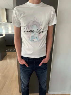 Vit Essence Style Cygne T-shirt - Fräsch vit t-shirt från Essence Style med tryck av en svan och en pampig portal på bröstet. Modellen är kortärmad och har rund hals. Tillverkad i mjuk Supima bomull som känns lyxig mot huden. Perfekt för dig som gillar stilrena prints och skandinavisk design.