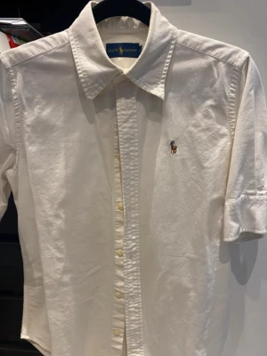 Vit skjorta från Ralph Lauren - Klassisk vit skjorta från Ralph Lauren med broderad logga på bröstet. Skjortan har kort  ärm, knappar framtill och en snygg krage. Tillverkad i bomull för en skön och stilren känsla. Perfekt till jeans eller chinos.