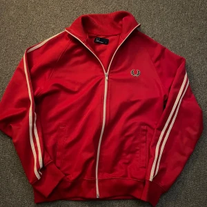 Fred Perry röd och svart ziptröja S - Två klassiska Fred Perry ziptröjor i storlek S. En röd och en svart, båda med vita ränder längs ärmarna och broderad logga på bröstet. Tröjorna har hög krage, muddar och dragkedja hela vägen. Perfekta för en sportig och retro look. 300kr styck eller båda för 500kr.