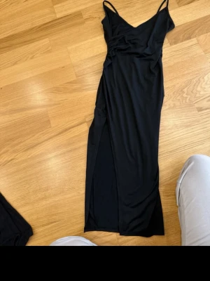Svart midiklänning med slits - Säljer en svart midiklänning med tunna axelband och v-ringad urringning. Klänningen har en snygg slits på sidan och är figurnära i modellen. Perfekt för dig som gillar minimalistisk och stilren look.