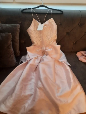 Ljusrosa långklänning från Monsoon - Elegant ljusrosa långklänning från Monsoon med tunna axelband, spetsdetaljer upptill och stor rosett i midjan. Kjolen är i glansigt satinliknande material och överdelen har blommig spets. Perfekt för speciella tillfällen.