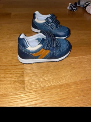 Blå sneakers med kardborreband - Säljer ett par blå sneakers för barn med vita detaljer och två orangea ränder på sidan. Skorna har kardborreband för enkel på- och avtagning, vit sula och rund tå. Materialet är en mix av syntet och textil, perfekt för lek och vardag. Aldrig använda 