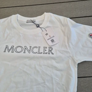 Vit Moncler t-shirt med logga - Snygg vit t-shirt från Moncler i bomull med stor logotyp på bröstet och ett broderat Moncler-märke på ärmen. Klassisk rund hals och korta ärmar. Perfekt till jeans eller shorts för en clean och stilren look.Liten i storlek.