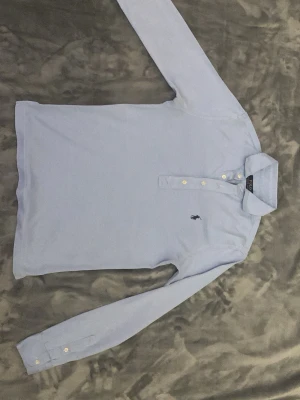 Ljusblå långärmad pikétröja Polo Ralph Lauren - Säljer en klassisk ljusblå långärmad pikétröja från Polo Ralph Lauren i 100% catton. Tröjan har knappar framtill, krage och den ikoniska broderade loggan på bröstet. Perfekt för en clean och stilren look. den är fortfarande lämplig för XS-S.