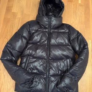 Svart pufferjacka från RYVLS - Säljer en svart glansig varm pufferjacka från RYVLS med huva och dragkedja framtill. Storlek 170/76 men är som motsvarande XS/S. Sparsamt använd. 