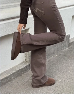 Bruna bootcut jeans från Nelly  - Säljer ett par bruna bootcut jeans från Nelly med snygga kontrastsömmar och ikonisk zigzag-detalj på bakfickorna. Jeansen har låg midja och är tillverkade i mjuk bomull med lite stretch för skön passform. Perfekta till sneakers eller boots. Har använts ett få tal gånger☺️