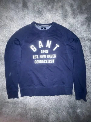 Vintage  sweatshirt från GANT - Säljer en marinblå sweatshirt från GANT med vit text framtill: 'GANT 1949 EST. NEW HAVEN CONNECTICUT'. Tröjan har rund halsringning, ribbade muddar och är långärmad. Klassisk och enkel design i mjuk bomull.