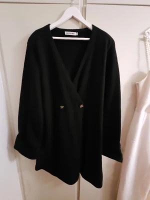 Svart omlottklänning från Adoore - Säljer en svart omlottklänning från Adoore med v-ringning och dekorativa knappar framtill. Klänningen har lång ärm och en stilren, minimalistisk look som passar perfekt till många olika tillfällen.