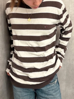 Tommy Hilfiger - randig tröja  - Säljer en brun och vit randig tröja från Tommy Hilfiger med klassisk rund hals och diskret logga på bröstet. Tröjan har långa ärmar och är gjord i mjuk bomull, perfekt för en chill och avslappnad stil.