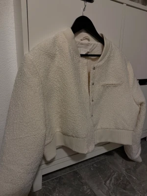 Vit teddy bomberjacka med tryckknappar - Säljer en supermjuk och fluffig vit teddy bomberjacka med croppad passform. Jackan har ribbade muddar, tryckknappar framtill och en liten bröstficka som detalj. Perfekt för dig som vill ha en mysig och trendig look till hösten och våren.