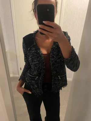 Svartvit cardigan jacka med dragkedjor - Säljer en svartvit cardigan jacka med fransiga kanter och coola dragkedjor vid fickorna. Jackan är stickad med inslag av vitt och svart, har lång ärm och en lite croppad passform. Perfekt för dig som gillar en trendig och unik look.