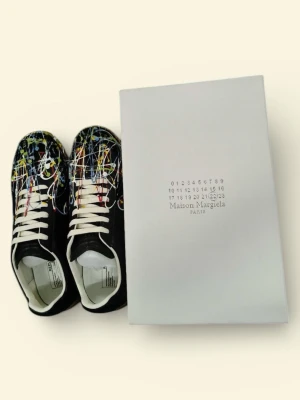 Maison Margiela sneakers med färgstänk - Unika svarta sneakers från Maison Margiela med färgstänk i vitt, blått, gult och rött. Skorna har vit snörning, beige gummisula och är tillverkade i skinn. Perfekt för dig som vill sticka ut med en konstnärlig vibe.