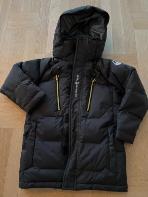 Svart gåsdun jacka i Gore-Tex från Sail Racing, jr - Säljer en svart/antracitgrå gåsdun jacka från Sail Racing med huva och två gula dragkedjor framtill. Jackan är i Gore-Tex och har lång ärm, quiltad design och justerbara muddar. Perfekt för kalla dagar, är i stl 120 cl ca 6-7 år. Jackan är helt ny och helt oanvänd. 