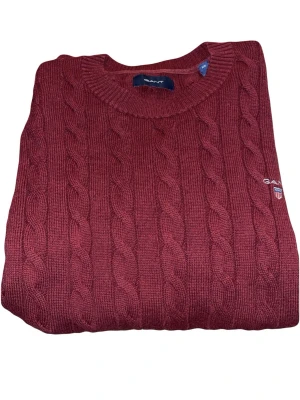 Vinröd kabelstickad tröja från GANT - Vinröd kabelstickad tröja från GANT med rund halsringning och broderad logga på bröstet. Tröjan har klassisk kabelstickad struktur och ribbade muddar vid ärmslut och nederkant. 