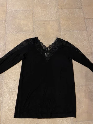 Svart spetströja från Culture - Svart långärmad tröja från Culture med v-ringning och breda spetsdetaljer längs halsringningen och axlarna. Toppens material känns mjukt och följsamt, och spetsen ger en snygg och elegant touch.