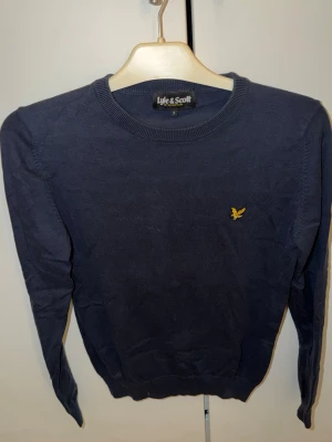 Marinblå tröja - Lyle & Scott - Snygg marinblå tröja från Lyle & Scott i storlek S. Klassisk rund hals och långärmad modell med gul broderad logga på bröstet. Perfekt för dig som gillar stilrena och enkla plagg. Materialet är mjukt och skönt bomull.