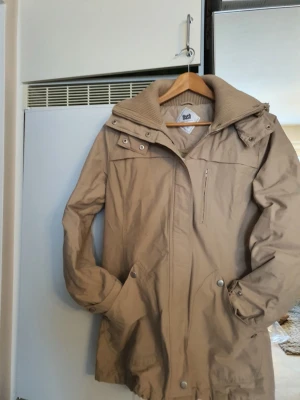 Beige jacka från Flash - Säljer en beige jacka från Flash med huva och hög krage. Jackan har dragkedja och tryckknappar framtill, samt praktiska fickor med knappstängning. Materialet är 70% bomull och 30% nylon, vilket gör den både slitstark och bekväm. Perfekt för kyligare dagar.