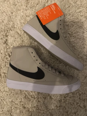 Nike Blazer Mid 77 HF sneakers - Snygga Nike Blazer Mid 77 HF sneakers i ljusgrått med svart swoosh och vit sula. Klassisk hög modell med snörning och vadderad plös. Ovandelen är i syntetmaterial och har en stilren look som passar till många outfits.