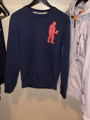 Mörkblå Off-White sweatshirt S - Säljer en mörkblå sweatshirt från Off-White i storlek S. Tröjan har ett stort neonrött tryck av en figur på både fram- och baksidan samt vit Off-White-logga på ryggen. Tillverkad i mjuk bomull och har långa ärmar och ribbade muddar.