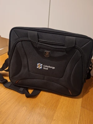 Svart datorväska Port Designs - Svart datorväska från Port Designs med Göteborgs Stad-logga. Väskan har två stora fack med dragkedja, vadderade handtag och justerbar axelrem. Materialet är slitstark polyester och insidan är ljusgrå. Perfekt för laptop och dokument.