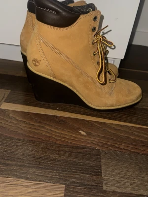 Timberland boots med kilklack - Snygga Timberland boots i beige mocka med mörkbrunt vadderat skaft och klassiska gula snören. Skorna har hög kilklack i svart och rund tå. Perfekta för dig som vill ha en robust men ändå trendig look. Timberland-logga på sidan.