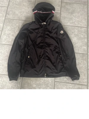 Svart vindjacka från Moncler - Snygg svart vindjacka från Moncler med huva och dragsko. Jackan har dragkedja framtill, två fickor med tryckknappar och Moncler-logga på ärmen. Huvan har en vit och röd rand som detalj. Perfekt för dig som vill ha en stilren och sportig look.