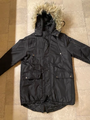 Svart parkajacka med fuskpäls - Svart parkajacka med huva och fuskpäls, flera fickor med knappar och dragkedja framtill. Jackan har lång ärm och quiltad insida för extra värme. Perfekt för kalla dagar och har en klassisk, stilren look.