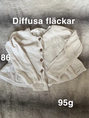 Vit kofta från Newbie med knappar - Söt vit kofta från Newbie i storlek 86. Koftan har lång ärm, rund hals och fem träknappar framtill. Perfekt att slänga över en t-shirt för extra värme.🌸Diffusa fläckar. Storlek 86