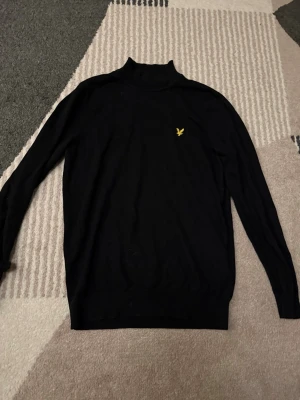 Svart polotröja från Lyle & Scott - Svart polotröja från Lyle & Scott med klassisk gul logga på bröstet. Tröjan har lång ärm och är tillverkad i mjukt material som passar perfekt för kyligare dagar. Enkel och stilren design som funkar till det mesta.