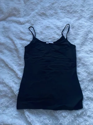 Svart linnetopp från ZARA - Säljer en enkel och stilren svart linnetopp från ZARA med tunna axelband. Toppen har en rak passform och är tillverkad i mjukt material som känns skönt mot huden. Perfekt att matcha med jeans eller kjol för en clean look. Den är lite noprig under armhålan