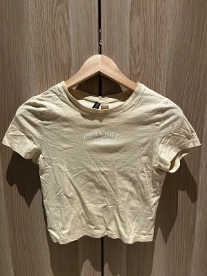 Beige kortärmad t-shirt Club Society - Snygg beige t-shirt från H&M med trycket 'CLUB SOCIETY MANCHESTER' på bröstet. Modellen är croppad med ribbad rund halsringning och korta ärmar. Perfekt till jeans eller kjol för en chill look.