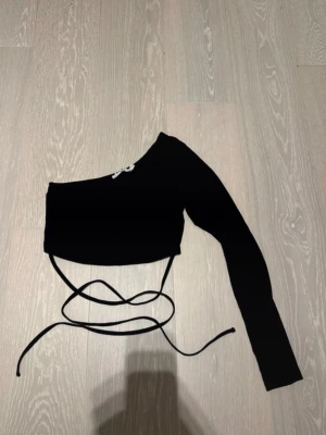 Svart oneshoulder topp med snörning - Trendig svart croppad topp med oneshoulder-design och lång ärm från NAKD. Toppen har smala knytband i midjan! Använd en gång!