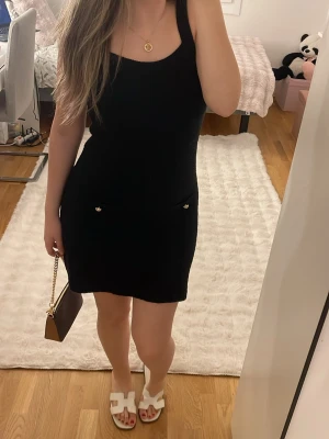 Svart stickad kortklänning från H&M - Supersnygg svart stickad klänning med breda axelband och två små fickor framtill dekorerade med guldfärgade knappar. Klänningen har en tight passform och är ärmlös, perfekt för en trendig look.