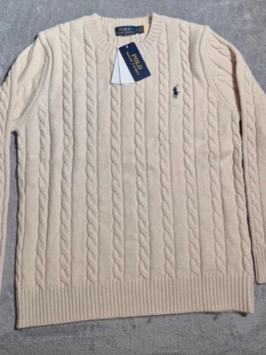 Beige kabelstickad tröja från Polo Ralph Lauren - Kabelstickad tröja i beige från Polo Ralph Lauren med rund halsringning och broderad marinblå logga på bröstet. Tröjan har ribbade muddar vid ärmslut och nederkant, och är tillverkad i mjukt material som passar perfekt för kyligare dagar.