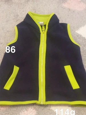 Mörkblå fleeceväst med lime detaljer - Säljer en mörkblå fleeceväst med limegröna detaljer och dragkedja framtill. Västen har två fickor med limegröna kanter och matchande kantband runt ärmhål och hals. Perfekt för lager-på-lager under kyliga dagar.🌸 Fint använt skick. Storlek 86