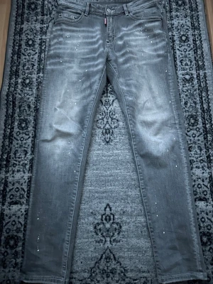 Grå Dsquared2 jeans - Säljer ett par grå jeans från Dsquared2 med coola vita färgstänk och slitningar för en edgy look. Klassisk femficksmodell med Dsquared2-tag på fickan och stor röd CATEN HEATED-patch bak. Raka ben och normal passform, perfekta till streetwear-stilen.