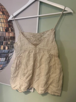 blus från zara - Säljer en söt beige linnetopp med tunna axelband och v-ringad hals. Toppen har broderade blommor upptill och fina spetsdetaljer längs sömmarna. Materialet är linne och toppen har en loose passform som ger en avslappnad vibe.