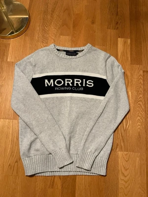 Grå stickad tröja Morris Rowing Club - Snygg grå stickad tröja från Morris Rowing Club med svart och vit panel över bröstet och tydlig logotyp. Tröjan har rund hals, ribbade muddar och lång ärm. Perfekt för dig som gillar klassisk stil med sportig touch.