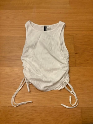 Vit ribbad linnetopp från H&M Divided, storlek S - Säljer en vit ribbad linnetopp från H&M Divided i storlek S. Toppen har snörning i sidorna som gör att du kan justera längden och skapa en snygg rynkad effekt. Ärmlös och i mjukt stretchigt material, perfekt till sommaren.